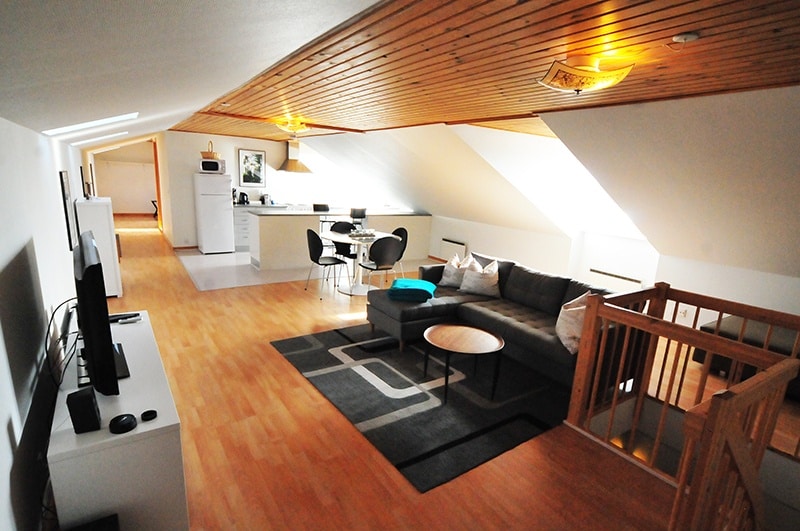 En moderne loftslejlighed med træloft og trægulv, opholdsstue med grå sektionssofa, spisebord med stole og køkken udstyret med hårde hvidevarer. Trapper er synlige til højre.