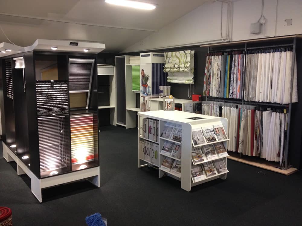 Interiør i en butik, der viser forskellige vinduesbehandlinger, herunder persienner, gardiner og gardiner. Prøvebøger og kataloger er placeret på et centralt bord, og hylder langs væggene.