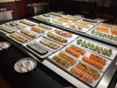 En buffet-udstilling viser flere fade med sushi-varianter arrangeret pænt på en disk med tomme tallerkener og serveringsredskaber i forgrunden.