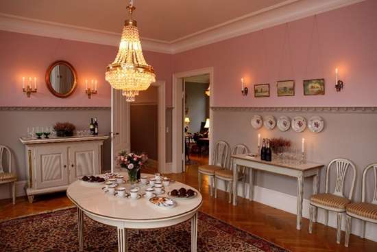Elegant spisestue med lysekrone, lyserøde vægge og vintagemøbler. Et hvidt bord er dækket med tekopper, underkopper og desserter. Tændt stearinlys pryder et sidebord, med indrammede billeder hængende på væggen.