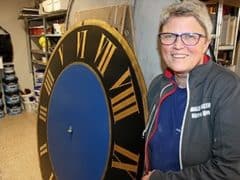 En person med kort gråt hår og briller står ved siden af en stor urskive med romertal på et værksted.
