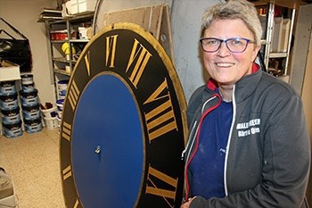 En person med kort gråt hår og briller står ved siden af en stor urskive med romertal på et værksted.