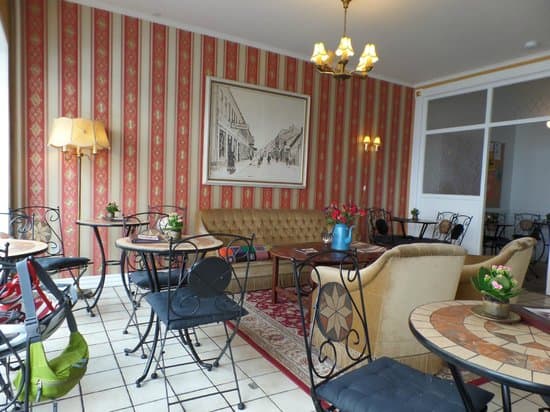 Et hyggeligt caféinteriør med en blanding af smedejern og polstrede siddepladser, mønstret tapet, et stort indrammet billede og flere små borde med blomsterarrangementer.