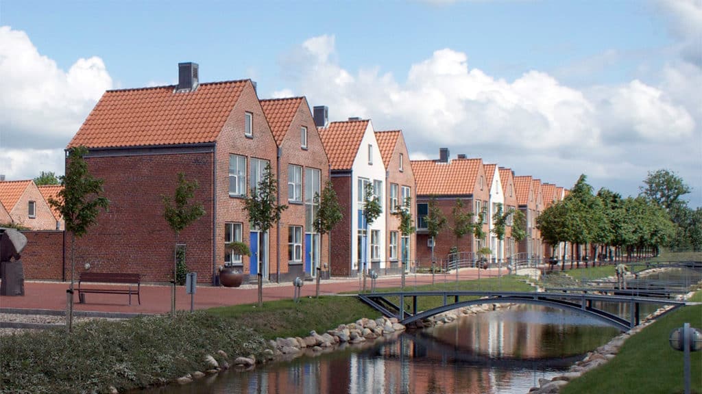 En række murstenshuse med orange tage, træer og en lille bro over en kanal på en klar dag.