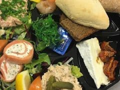 Et fad med forskellige fødevarer, herunder rundstykker, ost, rejer med mango, salat, lakseskiver, en syltet agurk og citronbåde.