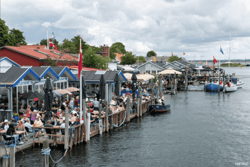 Overfyldt havnefront med folk, der sidder uden for restauranter, både, der ligger til kaj ved moler, og en overskyet himmel over hovedet.
