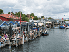 Overfyldt havnefront med folk, der sidder uden for restauranter, både, der ligger til kaj ved moler, og en overskyet himmel over hovedet.