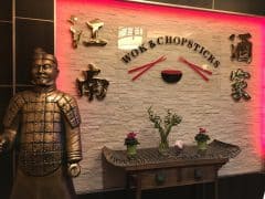 Statue af en kriger står ved et træbord med dekorative planter under et skilt, hvor der står "Wok & Chopsticks" i en restaurant dekoreret med kinesiske tegn og en lanterne.