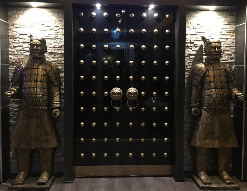 En sort dør med guldnitter er flankeret af to statuer af kinesiske krigere i bronzepanser, der står op ad en mur med stenfliser.