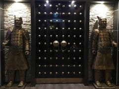 En sort dør med guldnitter er flankeret af to statuer af kinesiske krigere i bronzepanser, der står op ad en mur med stenfliser.