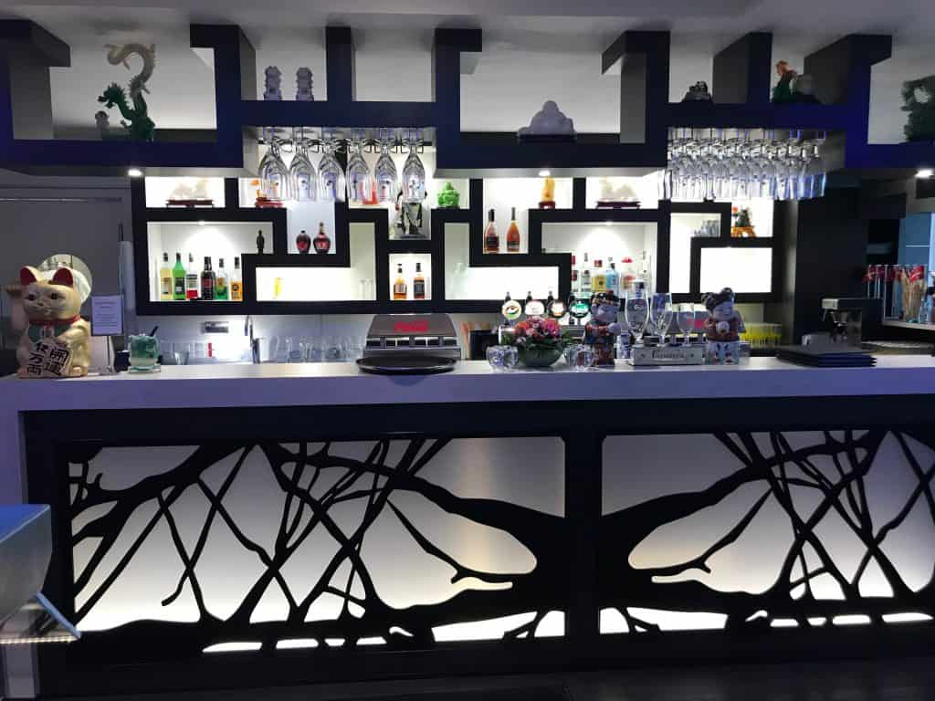 En moderne bar med et geometrisk, sort-hvidt design, forskellige flasker alkohol på oplyste hylder, glasvarer og eklektiske dekorationer, inklusive en Maneki-neko kattestatue.