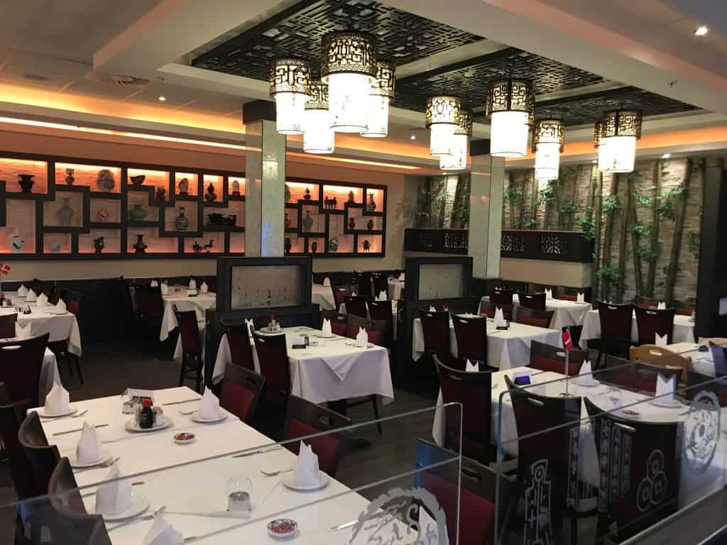 Et veloplyst, moderne restaurantinteriør med pænt arrangerede borde dækket af hvide duge, røde stole og dekorative lanterner hængende fra loftet. Hylder med små artefakter pryder væggene.