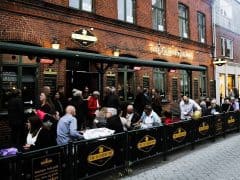 Et travlt udendørs opholdsområde på en murstenspub ved navn "The Oliver Plunkett" med mange gæster, der spiser og taler ved borde under aftenhimlen.