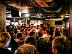 En fyldt barscene med folk, der står og snakker. Barområdet er godt oplyst med en række skilte og bannere over hovedet. Flasker og glasvarer er synlige på hylderne bag baren.