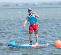 Byliv.nu: En person iført redningsvest og kasket padler, mens han står på et blåt og rødt paddleboard i roligt vand nær en orange bøje.