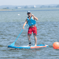 Byliv.nu: En person iført redningsvest og kasket padler, mens han står på et blåt og rødt paddleboard i roligt vand nær en orange bøje.