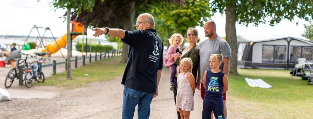 Byliv.nu: En mand peger, mens han taler med en gruppe på fem personer, heraf tre børn, på en udendørs park med legeredskaber og telte i baggrunden.