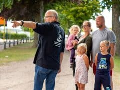 Byliv.nu: En mand peger, mens han taler med en gruppe på fem personer, heraf tre børn, på en udendørs park med legeredskaber og telte i baggrunden.