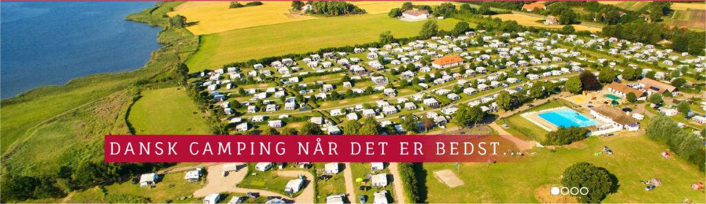 Byliv.nu: Luftfoto af en stor campingplads med talrige telte og mobile homes nær en vandmasse og grønne marker, med sætningen