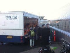 Byliv.nu: Folk er samlet på et mobilt fiskemarked, parkeret ved siden af en bygning. På siden af lastbilen står der "FRISK FISK FRA EGEN KUTTER." En cykel er i forgrunden.