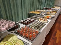 Byliv.nu: Et langt bord dækket med en hvid klud er fyldt med forskellige desserter, herunder kager, tærter og frugtkopper, alt sammen arrangeret på sølvbakker. Et grønt gardin fungerer som baggrund.