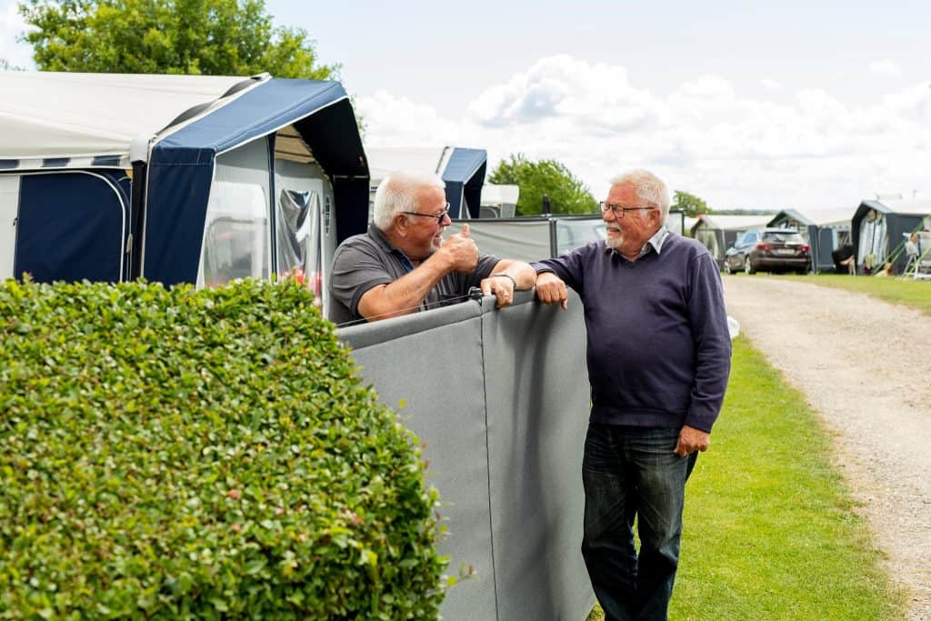 Byliv.nu: To ældre mænd taler muntert over et lavt hegn på en campingplads med telte, biler og frodigt grønt synligt i baggrunden.