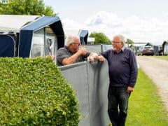 Byliv.nu: To ældre mænd taler muntert over et lavt hegn på en campingplads med telte, biler og frodigt grønt synligt i baggrunden.
