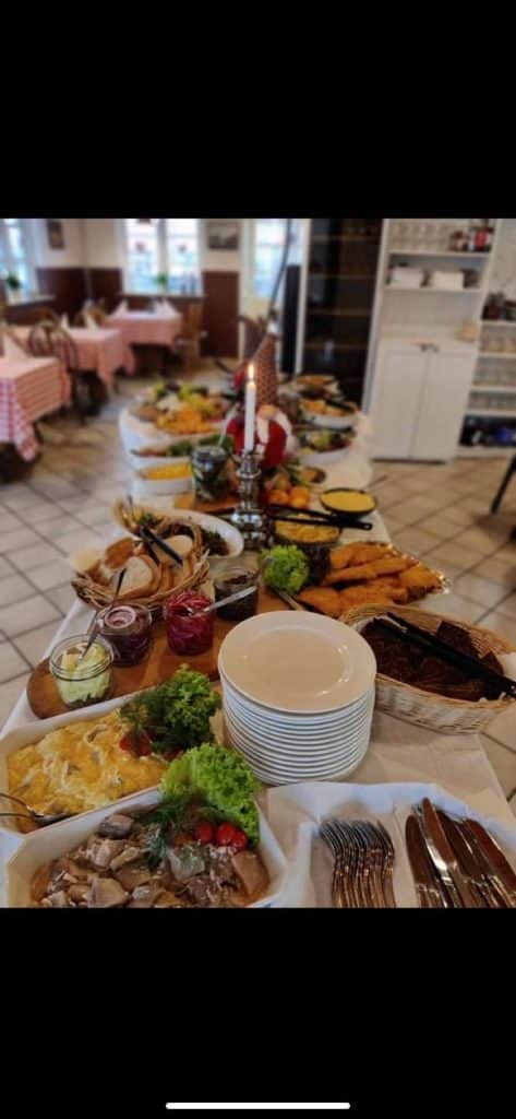 Byliv.nu: Et langt buffetbord er dækket med forskellige retter, herunder salater, kød, tallerkener og redskaber i en spiseplads med ternede duge og et stearinlys i midten.