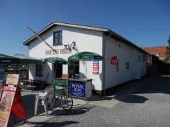 Byliv.nu: En hvid bygning med et lille udendørs opholdsområde med grønne paraplyer og stole. Skilte annoncerer "Royal Beer", "Tuborg" og is. En cykel er parkeret ved indgangen. På skiltet står der "Martens Røgeri.
