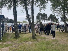 Byliv.nu: En gruppe mennesker står og taler i et parkområde med træer og parkerede køretøjer i baggrunden. Nogle er i nærheden af et picnicbord, og nogle få holder cykler.