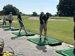 Byliv.nu: Folk øver deres golfsving på en driving range med spande med golfbolde i nærheden og et grønt landskab i baggrunden.