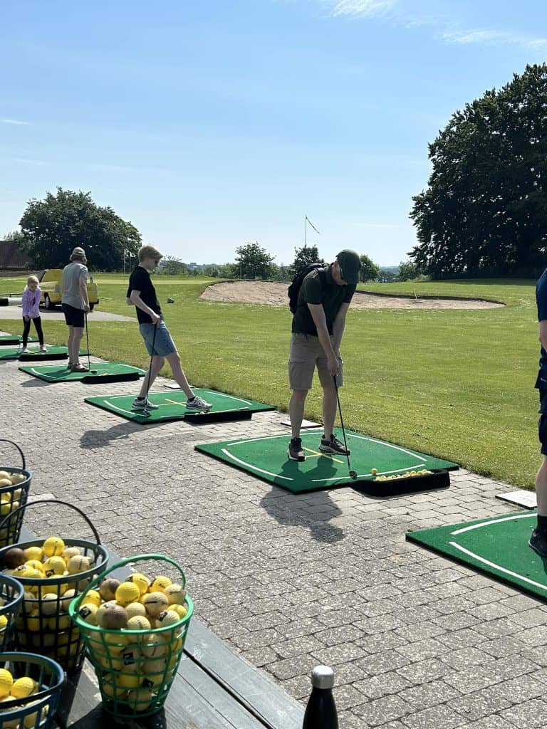 Byliv.nu: Folk øver deres golfsving på en driving range med spande med golfbolde i nærheden og et grønt landskab i baggrunden.