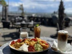 Byliv.nu: En tallerken nachos med toppings og tre drikkevarer på et bord på en udendørs café med palmer og en naturskøn udsigt i baggrunden.