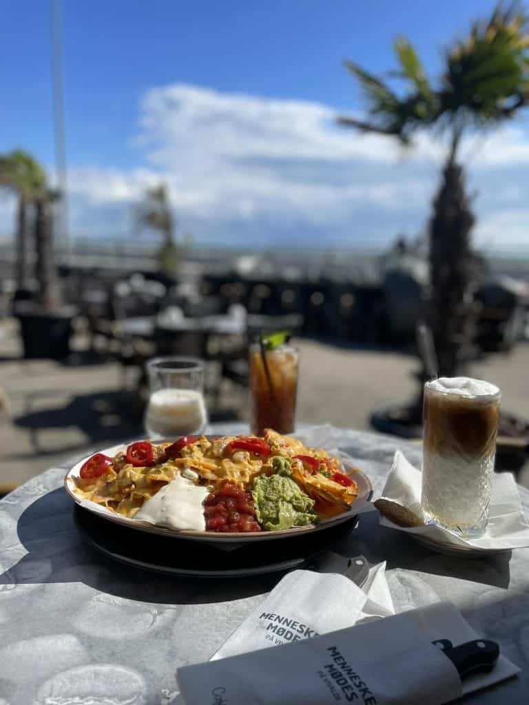 Byliv.nu: En tallerken nachos med toppings og tre drikkevarer på et bord på en udendørs café med palmer og en naturskøn udsigt i baggrunden.