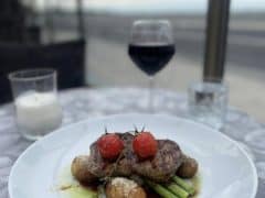 Byliv.nu: En ret bestående af bøf, cherrytomater, kartofler og asparges står på et marmorbord ved havet, ledsaget af glas rødvin og vand og et tændt stearinlys.