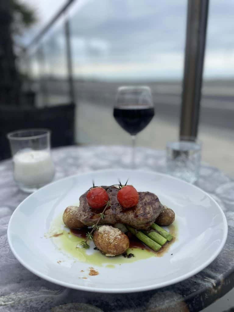 Byliv.nu: En ret bestående af bøf, cherrytomater, kartofler og asparges står på et marmorbord ved havet, ledsaget af glas rødvin og vand og et tændt stearinlys.