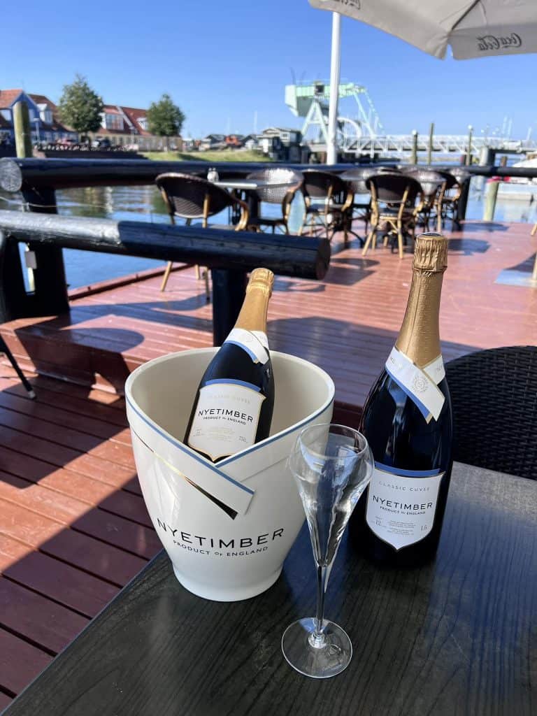 Byliv.nu: To flasker Nyetimber mousserende vin, den ene i en spand med is og den anden på et bord ved siden af en champagnefløjte. Indstillingen er et udendørs dæk ved vandet med ekstra siddepladser i baggrunden.