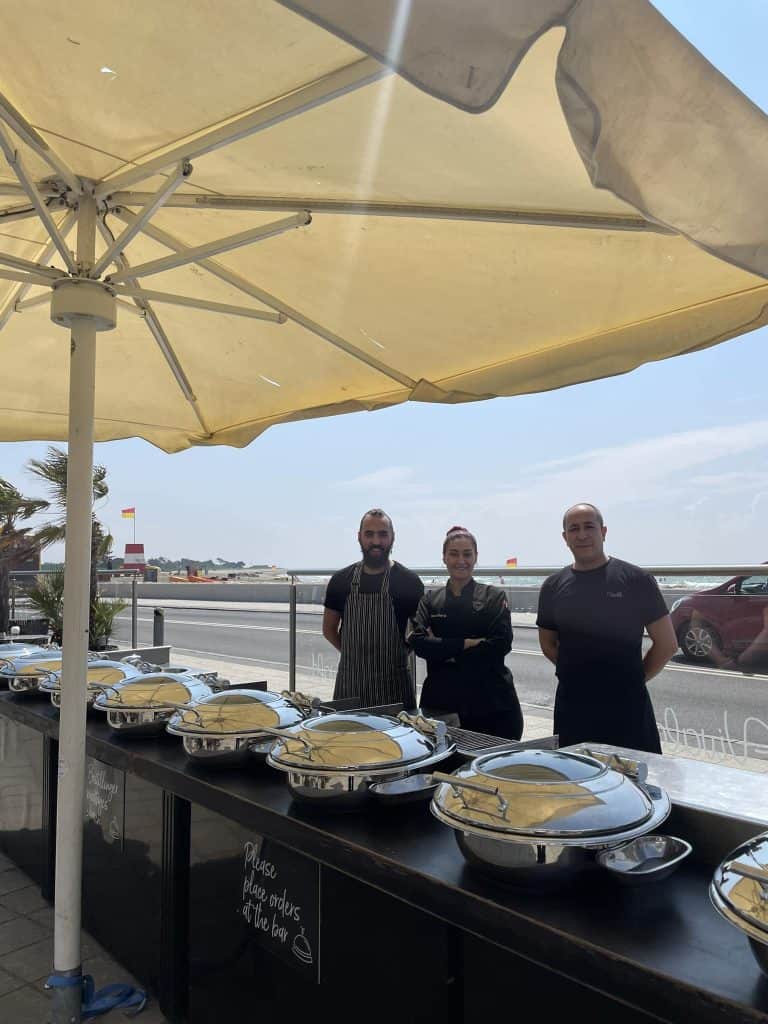 Byliv.nu: Tre personer står bag et buffetbord under en stor terrasseparasol, med gnavende fade klar til servering. En indstilling ved stranden er synlig i baggrunden.