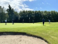 Byliv.nu: Folk er på en golfbane og putter i nærheden af hullet, med en golfspiller bøjet over og forbereder sig på at putte. Træer og en sandbunker er synlige i baggrunden.