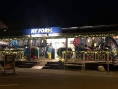 Byliv.nu: En butiksfacade mærket "NY FORM" viser forskellige jakker og tøj under lysslynge om natten. Et skilt og dekorativ slæde er synlig nær indgangen.