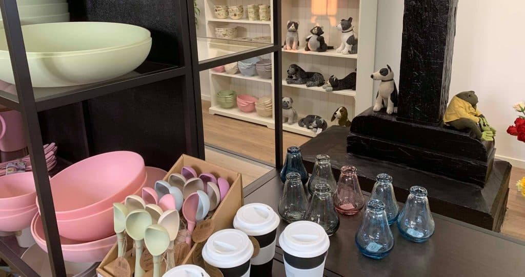 Byliv.nu: En butiksudstilling med pastelfarvede skåle, skeer og kopper med keramiske hundefigurer på hylder i baggrunden og glasvaser i forgrunden.