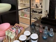 Byliv.nu: En butiksudstilling med pastelfarvede skåle, skeer og kopper med keramiske hundefigurer på hylder i baggrunden og glasvaser i forgrunden.