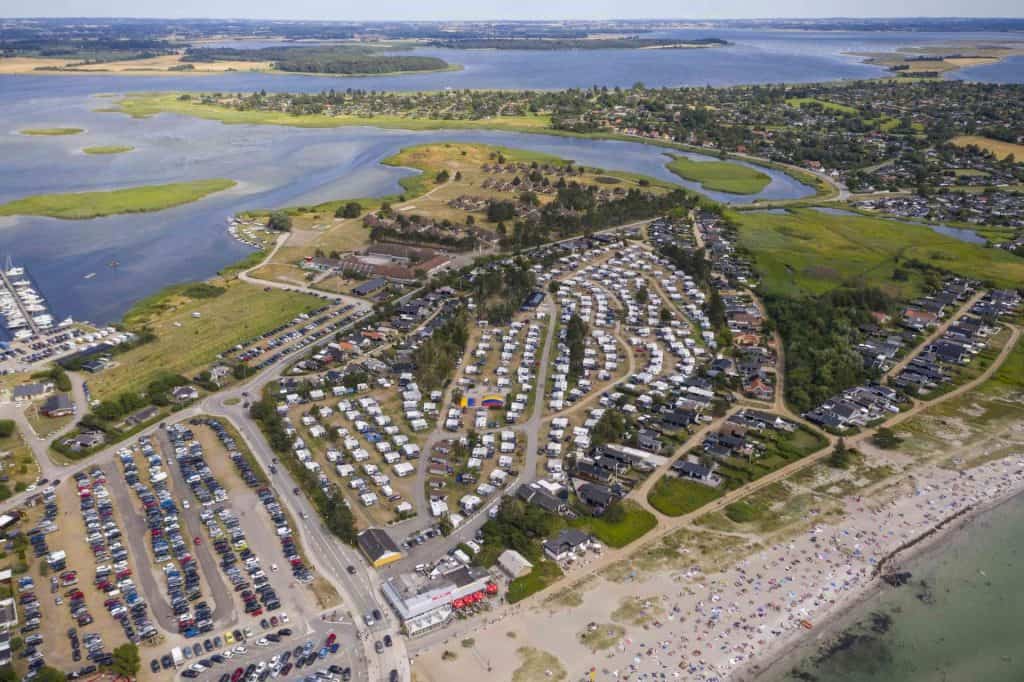 Byliv.nu: Luftfoto af et kystområde med en stor campingplads med talrige trailere, tilstødende parkeringspladser, en strand med rekreative aktiviteter og omkringliggende vandområder og grønne områder.