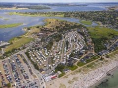 Byliv.nu: Luftfoto af et kystområde med en stor campingplads med talrige trailere, tilstødende parkeringspladser, en strand med rekreative aktiviteter og omkringliggende vandområder og grønne områder.