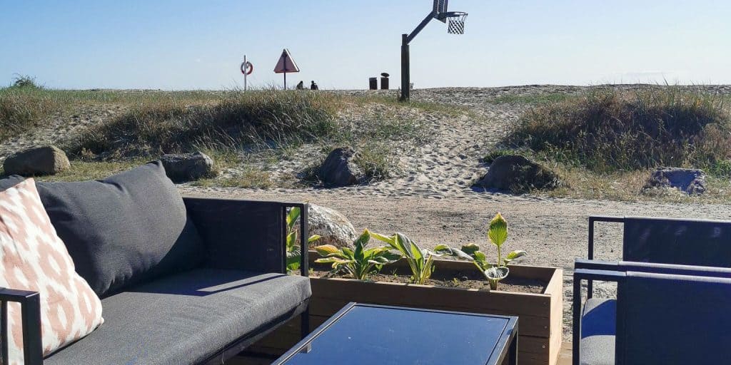 Byliv.nu: Udendørs opholdsområde med grå møbler og en lille plantekasse, med udsigt over en sandet sti, der fører til en strand. En basketballkurv er synlig i baggrunden nær nogle skilte og græs.