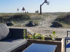 Byliv.nu: Udendørs opholdsområde med grå møbler og en lille plantekasse, med udsigt over en sandet sti, der fører til en strand. En basketballkurv er synlig i baggrunden nær nogle skilte og græs.
