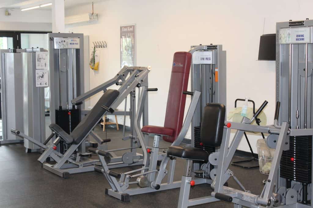 Byliv.nu: Et fitnesscenter med flere træningsmaskiner, herunder en brystpresse, en romaskine og en multifunktionsbænk. Rummet har et rent, organiseret udseende med lyse vægge.
