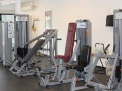 Byliv.nu: Et fitnesscenter med flere træningsmaskiner, herunder en brystpresse, en romaskine og en multifunktionsbænk. Rummet har et rent, organiseret udseende med lyse vægge.
