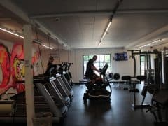 Byliv.nu: En mand træner på en ellipsemaskine i et fitnesscenter med løbebånd, vægtmaskiner og en væg med graffitikunst. En anden person er synlig på et løbebånd. Et TV er monteret på væggen.