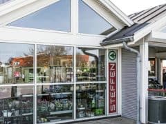 Byliv.nu: Udvendigt billede af et havecenter med et "Steffen"-skilt, store glasvinduer med planter indeni, en hvid bygningsstruktur og en overdækket indgang med mennesker og haveartikler.